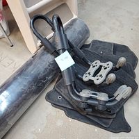 Photo of two black metal kayak racks with mounting hardware and several black floor mats for Subaru cars, along with a large black roll of weed barrier fabric.