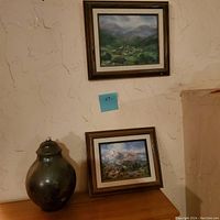 View of two framed oil paintings hung on textured wall with a glass vase placed below the lower painting.