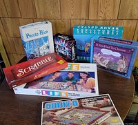 Photo showing seven board games arranged on a wooden table against a wood-paneled wall: Puerto Rico, Truth Bombs, Guesstures, If You Had To Choose..., Scrabble, The Game of Life, and Ginny-O.