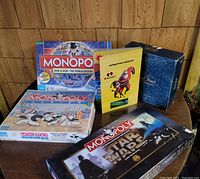 Four boxed board games and a puzzle game arranged on a wooden table against paneled wall: Monopoly Here & Now World Edition, Monopoly Junior, Monopoly Star Wars Edition, The Incredibles puzzle game.