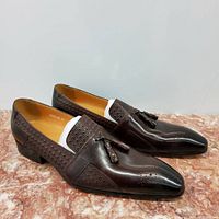 Pair of men's brown leather dress shoes with pointed toes, textured details, and tassels, photographed on a pink and white marbled surface against a white background.