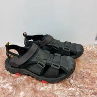 Pair of new men's black leather hiking sandals size 44 with closed-toe design and adjustable velcro straps, resting on a brown textured surface.
