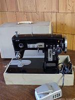 Front and side view showing the entire vintage Japanese sewing machine in black with attached power cord and foot pedal, placed on its carrying case.