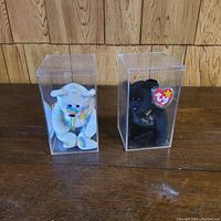 Photo of two Ty Beanie Babies bears in clear plastic display cases on a wooden surface against a wood paneled background. One bear is white with a rainbow tie, the other is black with 'The End Bear' embroidery and attached Ty tags visible.