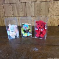 Three TY Beanie Babies in clear plastic display cases placed on wooden table against wood panel wall.