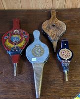 Four vintage fireplace bellows arranged on a wooden surface, showing different decorative styles and materials