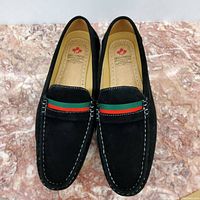 Top-down view of pair of black suede loafers showing white stitching and green/red strap detail