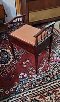 Photo of the vintage wooden piano stool showing the cushioned seat, wooden armrests with spindle supports, and tapered legs on a patterned carpet.
