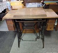 Front view of antique oak Singer sewing machine table featuring cast iron treadle base and two small drawers, with wooden top and fold-out extension panels stacked on top.
