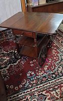 Wooden tea cart shown with drop leaf sides open, showcasing top surface, drawer, and lower shelf with large wood spoked wheels visible on side.