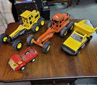 Four Tonka toy vehicles on wooden table showing front and side views including motor graders, dump truck, and pickup truck.