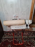 Side view of the Easy Ironer with beige fabric covered roller on red stand against wood panel wall