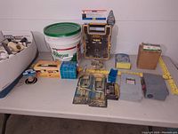 Wide shot of assorted plumbing fittings in a box alongside drywall compound bucket, tile cutter, small electrical boxes, measuring tape, and small hardware