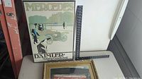 Framed Mercedes Daimler vintage poster featuring an early automobile and figures, alongside a ruler for scale.