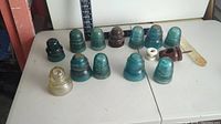 Thirteen vintage glass electrical insulators displayed on a table in two rows, showing various sizes and colors including blue, teal, brown, white, and clear glass.