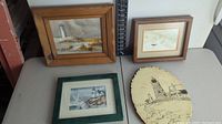 Photo of two framed lighthouse art pieces and one woodburning artwork on a wooden slab on a table.