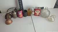 Overview of vintage oil cans, metal and glass containers including Nestle Hot Cocoa tin and decorative glass bottles set against a plain white background