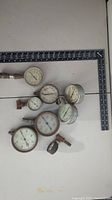 Seven old pressure gauges arranged on a flat surface with a black square ruler to show size comparison.