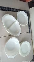 Plain white Pyrex dishes arranged on a white surface including an oval divided serving dish and oval scalloped plate.
