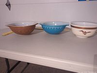 Side view of three Pyrex bowls showing different colors and patterns: brown with gold wheat, blue with white decoration, cream with eagle and chain design.