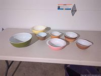 Full set of seven Pyrex bowls in various solid colors and sizes displayed on table with white wall background.