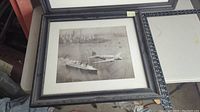 Two large framed prints showing transportation themes with ships and an airplane flying over a city coast. Frames are black with some ornamentation, prints are sepia tone. One frame back shown, paper backing partially damaged.