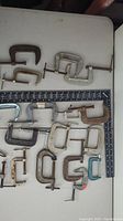Top view of various small C clamps arranged within a square measuring tool, showing different sizes and metal finishes including rust and silver tone.