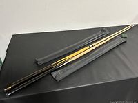 Two pool cues laid on a black surface, one in plastic protective wrapping and one unwrapped with parts separated.