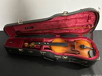 Violin inside open black hard case lined with red velvet with the violin bow secured in place.