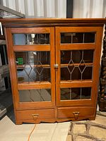 Front view of the antique wooden china cabinet showcasing two leaded glass doors and two bottom drawers with brass handles.