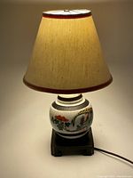 Lit view of the entire lamp showing the lampshade and painted vase base on wooden stand.