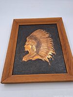 Framed copper portrait of a First Nations figure in profile with feather headdress against black background.