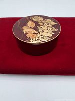 Image of the round wooden storage container lid featuring gold floral designs on a dark lacquer base, placed on a red cloth.