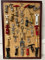 Framed corkscrew display with 29 assorted vintage corkscrews mounted on a cork board, showing a wide variety of handle types and finishes