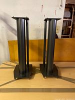 Front view of two black metal heavy-duty Mission speaker stands side by side on a wooden surface.