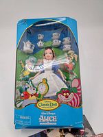 Front view of Alice in Wonderland doll in original blue box with tea set and backdrop graphics.