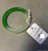 Jade bangle shown with a tag indicating 'Style: Emerald' and 'Size: Bracelet', highlighting the green and white coloring gradient and good condition.