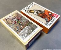 Photo of two tarot card boxes on a gray surface. One box is Tarot of A.E. Waite with the Fool card artwork on top. The other box is Paulina Tarot with ornate floral design.