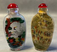 Both snuff bottles standing upright showing front detailed scenes and red stoppers.