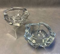 Two signed clear glass ashtrays, one hexagonal and one irregular scalloped shape, shown on clear stands and surface.