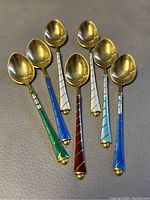 Seven vintage sterling silver teaspoons with colorful enamel handles arranged on a gray surface showing spoon bowls and handles.