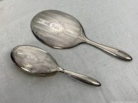 Both sterling silver hand mirror and hair brush shown from back with visible monogram and engraved linear and floral patterns.