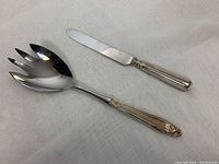 Pair of silver handled serving pieces on a white textured surface showing full length of knife and tong