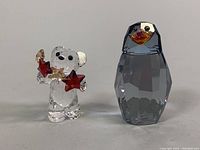 Front view of the clear crystal teddy bear holding stars and the smoked-colored penguin figurine standing side-by-side on a neutral background.
