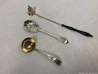 Full view of all three sterling silver items: a bell-shaped candle snuffer with black handle, a scalloped sugar spoon, and a round perforated tea strainer with gold-tone interior.