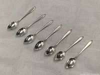 Seven sterling silver espresso spoons arranged in a row on a textured white cloth. Spoons have ornate handle ends and clean polished bowls.
