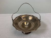 Front and top view of an old sterling silver pedestal candy dish with a scalloped edge and an attached handle, showing tarnishing on the surface.