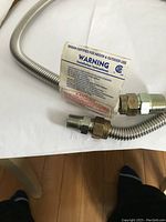 Close-up photo of gas appliance flex hose showing stainless steel corrugated hose with brass threaded connectors and warning label.