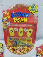 Front view of sealed Adroit Bean Game blister pack showing 4 yellow bean capsules with question mark designs, title, and promotional text.