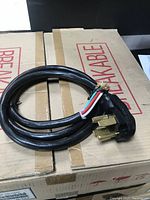Black 220 volt dryer cable coiled on cardboard box, showing plug with three brass prongs and four color-coded wire ends.
